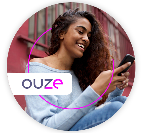 Mulher jovem, negra, sorrindo, sentada do lado de fora do prédio onde mora, olhando o celular 