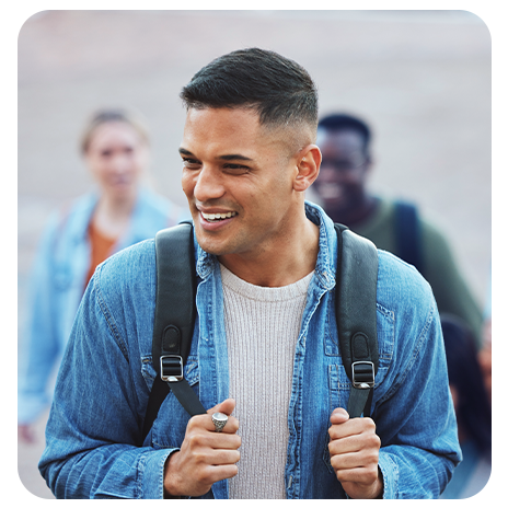 Homem Jovem, branco, sorrindo, de camisa jeans, mochila nas costas andando em um campus de faculdade.