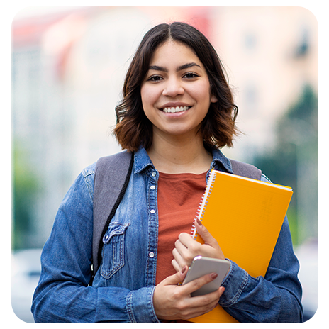 Mulher jovem, branca, de cabelos lisos e curtos, sorrindo, mochila nas costas, segurando um caderno e um celular. 