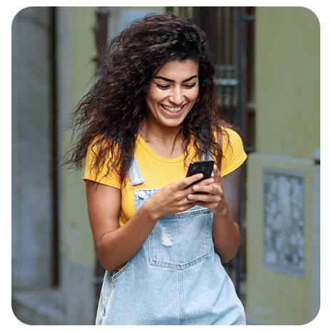 Mulher Jovem, branca, cabelos pretos, camiseta amarela, macacão jeans, sorrindo, mexendo no celular. 