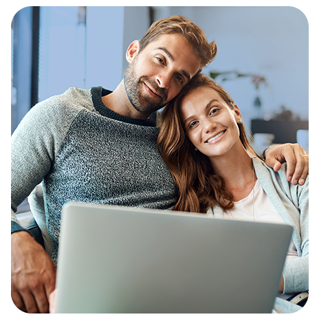 Casal, branco, abraçados, sorrindo, sentados em um sofá, com um notebook a frente deles.