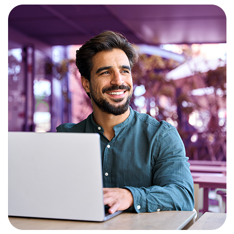 Homem jovem, branco, de barba, camisa azul, sorrindo, sentando em frente a uma mesa em um local externo, mexendo em seu notebook. 