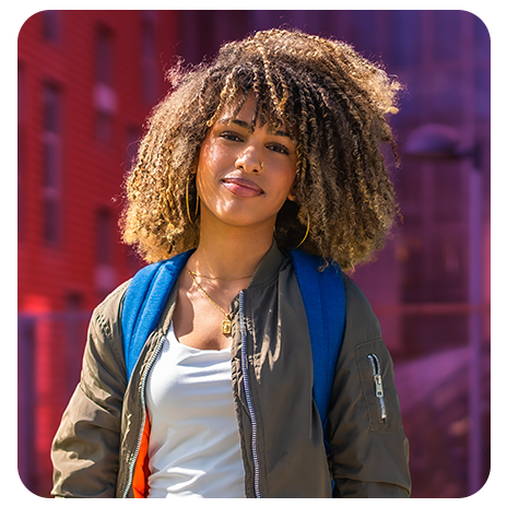 Jovem negra, sorrindo, de mochila, jaqueta marrom, camiseta branca.