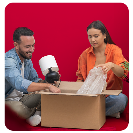 Casal sentado no chão abrindo uma caixa de mudança, retirando uma luminária e embalagens plásticas.
